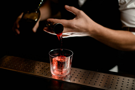 特写镜头。酒吧招待手拿着跳汰机，把红色的液体倒进加冰的玻璃杯里