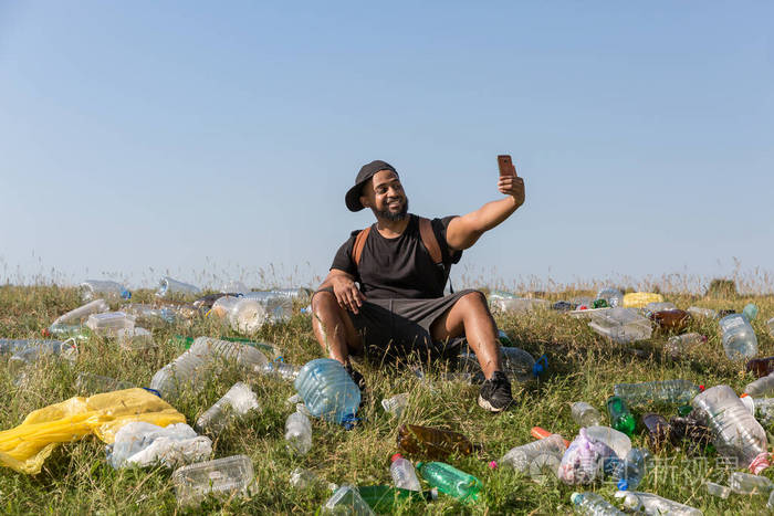 一个面带微笑的年轻人坐在一个绿色的斜坡上上面散落着塑料瓶
