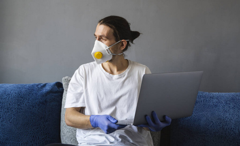 隔离期间，戴着医用面罩和橡胶手套的男子坐在家里，在沙发上用笔记本电脑工作。设计师艺术家建筑师商人在大流行性感冒的远程工作。