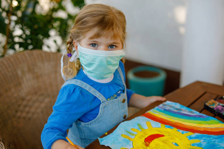 在大流行性冠状病毒检疫性疾病期间，戴着医用面罩的小女孩用水彩画彩虹。孩子们在世界各地画彩虹，上面写着让一切都好。