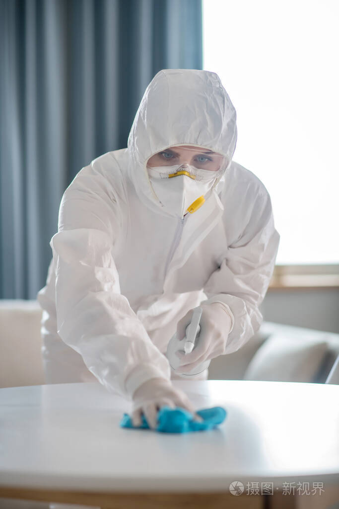 一位穿着白色工作服和防护手套的妇女在给桌子表面消毒时看起来很专注