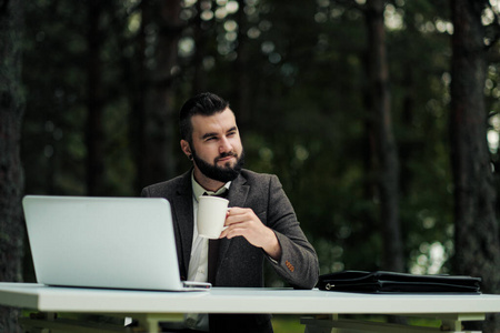 年轻迷人的商务男士西装革履，打着领带，坐在办公桌前，在户外工作。喝白杯子里的咖啡。背景是绿树自然和公园。