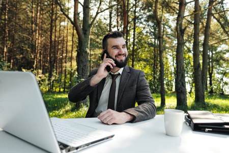 年轻迷人的商务男士西装革履，打着领带，坐在办公桌前，在户外工作。喝白咖啡，打电话。背景是绿树自然和公园。