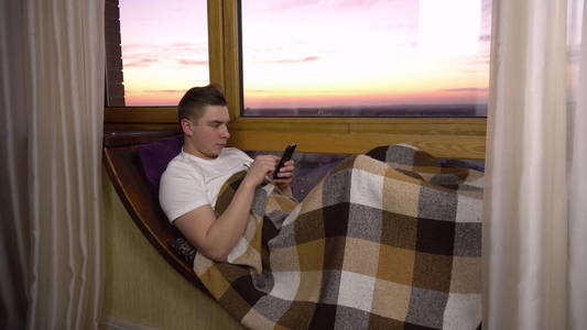 一个年轻人在电话里发短信。一名男子躺在靠窗的窗台上，手里拿着一部智能手机。晚上的窗外。