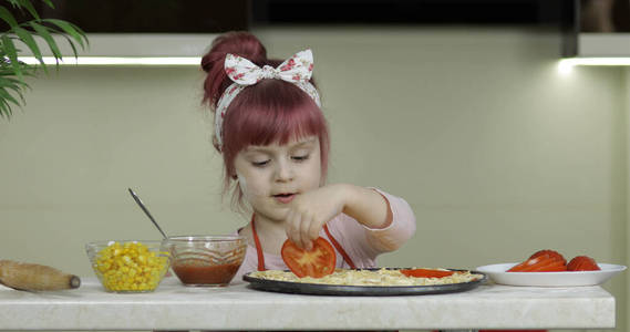 做披萨。厨房里围着围裙的小孩往面团里放西红柿片