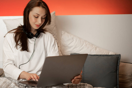 年轻漂亮的女孩戴着耳机坐在床上，在家里用笔记本电脑工作