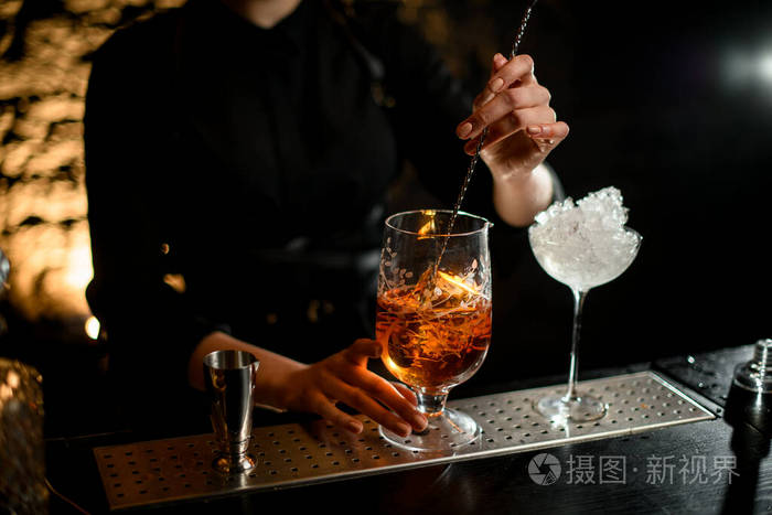 特写女调酒师用调羹在玻璃搅拌杯里混合冷鸡尾酒