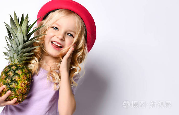 一个戴着红帽子和紫色上衣的金发小女孩。她微笑着，抚摸着她的脸，抱着菠萝，摆出一副孤立的白衣姿势