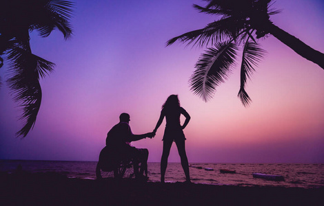 残疾人坐在轮椅上，妻子在沙滩上。日落时的剪影