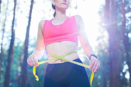 照片中，一位健康健康的年轻女士在森林里训练时，用卷尺测量腰围，控制体重