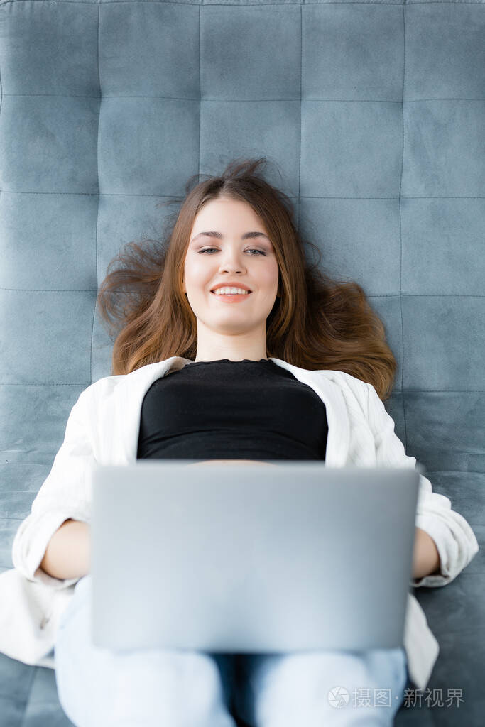 一个穿着白色夹克的女孩躺在沙发上，通过笔记本电脑工作，微笑着。在舒适的办公室工作。