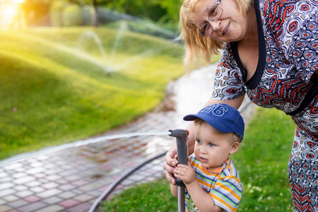 可爱可爱的白人金发男孩拿着水管洒水器和老奶奶在家后院浇灌花园。孩子小帮手和奶奶玩耍在夏季户外学习园艺