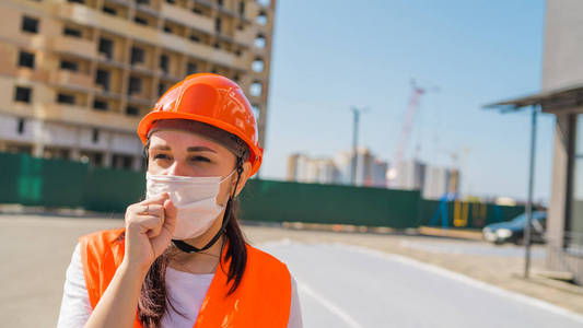 穿着工作服戴着医用口罩的女建筑工人在正在施工的房屋背景上咳嗽着拳头。冠状病毒流行性感染威胁的概念。