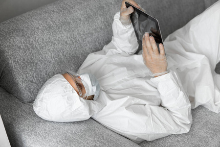 由于冠状病毒流行，身穿白色防护服和医用面罩的男子躺在沙发上工作。大流行期间的远程工作。在COVID19隔离期间呆在家里