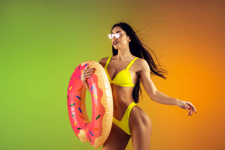 时尚写真年轻的健康和运动的妇女与橡胶甜甜圈在时尚的黄色泳装梯度背景。完美的身体为夏季做好准备。