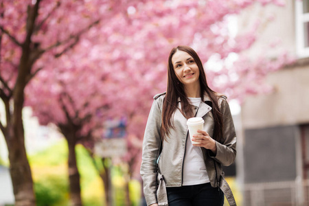 在樱花盛开的樱花树的背景下，一个穿着夹克和牛仔裤的黑发女孩在城市的户外