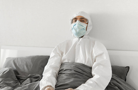 一个穿着白色防护服和面罩的人坐在卧室的床上等待冠状病毒隔离结束的画像。自我隔离。