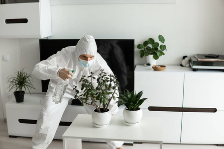 身穿防护服，戴着医用面罩和橡胶手套的男子在家里的客厅里用喷雾器照料着植物，而冠状病毒隔离剂COVID19。