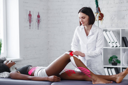 理疗师在患者身上应用运动机能学磁带膝盖。治疗师治疗年轻的美国黑人女运动员。运动机能学录音。创伤后康复运动理疗