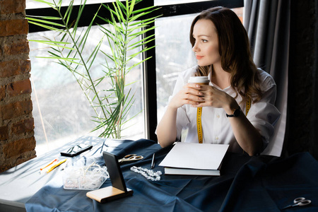 时装设计师休息了一会儿，手里拿着一个咖啡杯，坐在一张桌子旁，桌上摆着缝纫材料和一部智能手机。