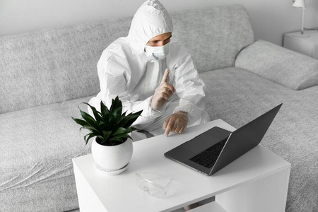 由于冠状病毒流行，身穿白色防护服和医用面罩的男子正拿着笔记本电脑在沙发上工作。大流行期间的远程工作。在COVID19隔离概念期
