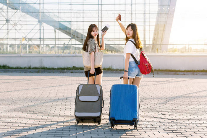 在机场候机楼附近，两位年轻迷人的亚洲女性穿着休闲服，提着行李，提着行李箱，手里拿着护照