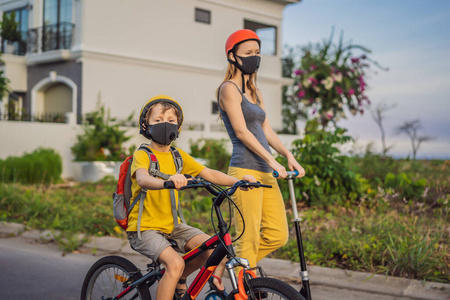 在阳光明媚的日子里，男孩和妈妈戴着医用面罩和安全帽骑着自行车背着背包。快乐的孩子在上学的路上骑自行车。你需要戴着面具上学因为
