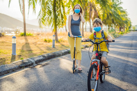 在阳光明媚的日子里，一个活跃的小男孩和他戴着医用面罩的妈妈骑着自行车。快乐的孩子在上学的路上骑自行车。因为冠状病毒流行，你得戴着