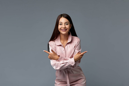 一个穿着粉红色工作服微笑的时尚女孩用手指指着空白的复印空间。复制空间，灰色背景。时尚理念。