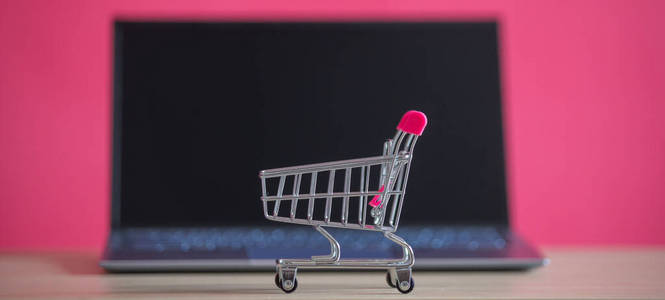 网上购物概念。粉红色背景的迷你购物车和笔记本电脑。小推车和个人电脑。