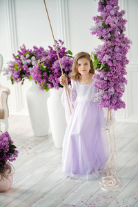 美丽的学龄前小女孩，穿着一件精致的连衣裙，坐在装饰着丁香花的秋千上
