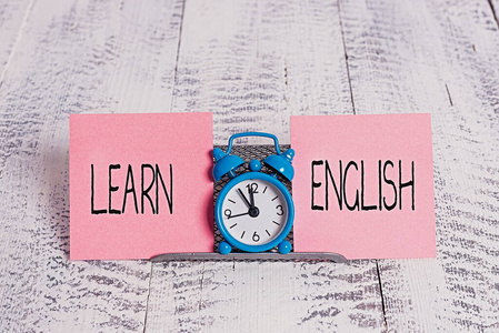 写笔记显示学习英语。商务照片展示通用语言，便于沟通和理解迷你蓝色闹钟站在两张纸之间的缓冲线上方。