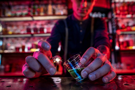 酒吧男招待准备好酒精鸡尾酒，在五颜六色的霓虹灯下点火喝，聚焦在玻璃杯上