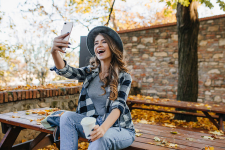 休闲装女士一头浅棕色头发的积极的女人在秋天公园喝咖啡自拍.