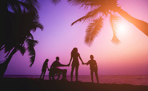 残疾人坐在轮椅上和家人在海滩上。日落时的剪影