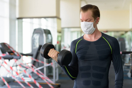 冠状病毒covid19感染期间戴口罩的年轻人在健身房用哑铃锻炼