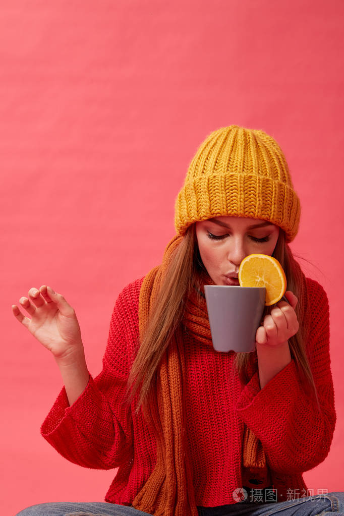 美丽的女孩穿着温暖舒适的毛衣拿着一杯橙汁的热饮料红色背景导播室