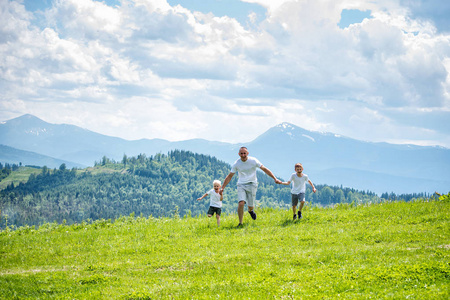 父亲和两个年幼的儿子手牵着手在绿色的田野上奔跑,背景是青山绿水