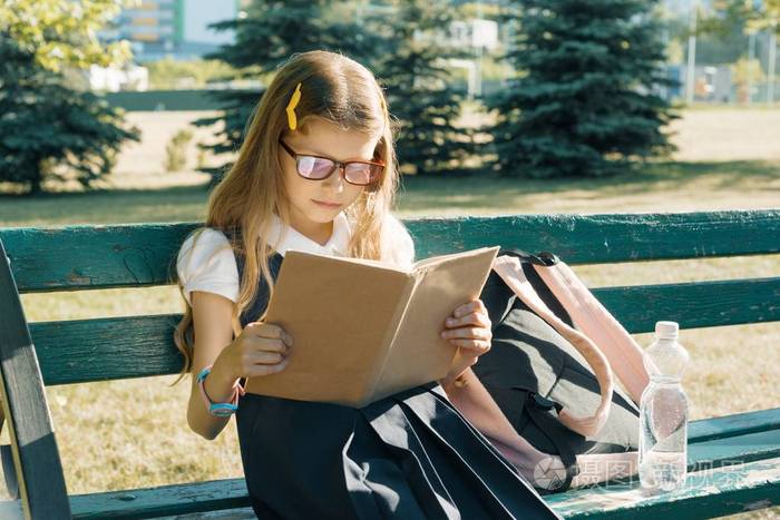 戴着眼镜的小女孩背着学校的书包坐在公园的长椅上看书
