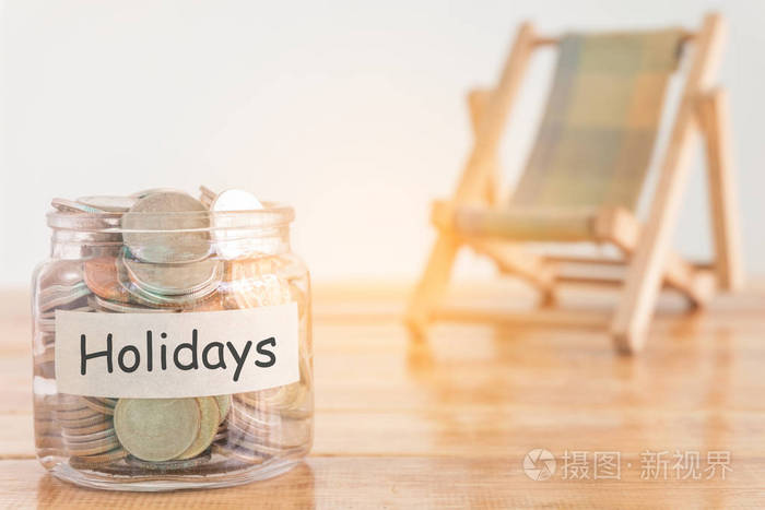 假期预算概念。假日储蓄概念。在钱罐里攒钱过节。木制背景上有硬币和沙滩椅的钱罐。