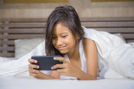 快乐美丽的7岁孩子在床上玩手机玩网络游戏,在年轻女孩和科技概念中
