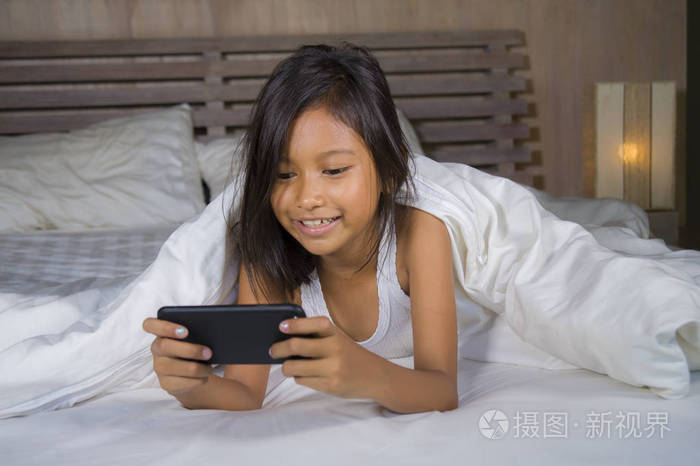 快乐美丽的7岁孩子在床上玩手机玩网络游戏在年轻女孩和科技概念中