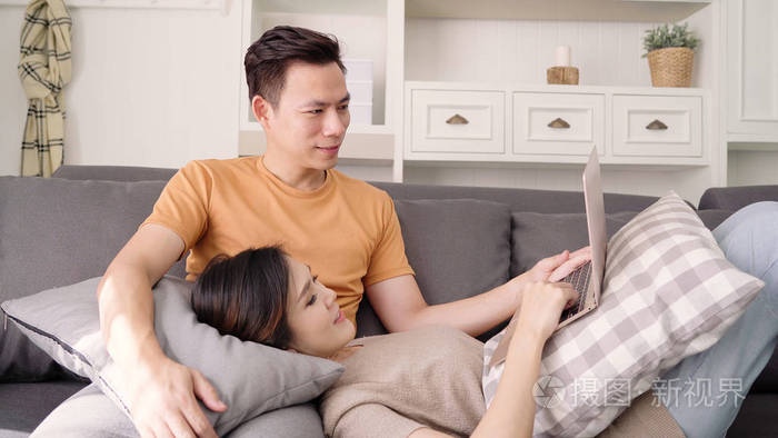 亚洲情侣在家里用笔记本电脑上网甜蜜的情侣在家休息时躺在沙发上享受