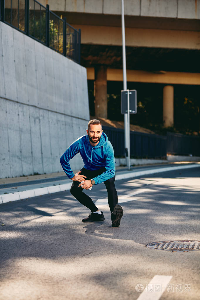 年轻的胡须男跑步者在跑步前伸展双腿穿着运动装健康的生活方式理念