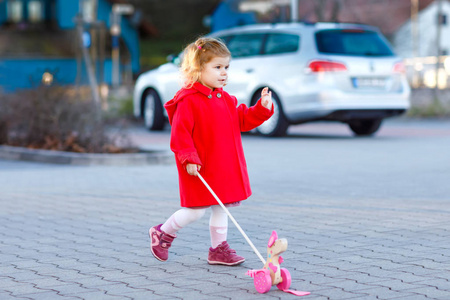 户外画像可爱的小女孩穿着红色外套在春天阳光明媚的日子推木玩具.