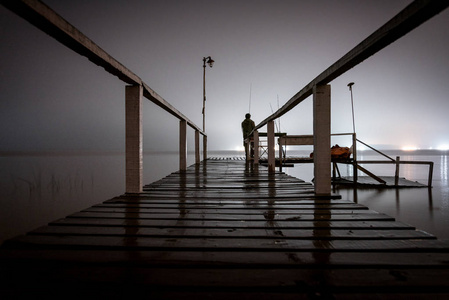 黑暗中站在木坞上的一个人的侧影,凝视着湖的空旷,湖水的另一面闪着