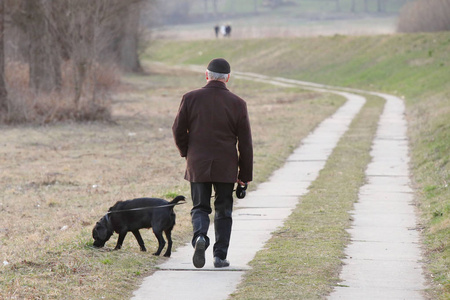 牵宠物一位穿着半季节服装的老人正牵着他的黑色拉布拉多犬在公园里