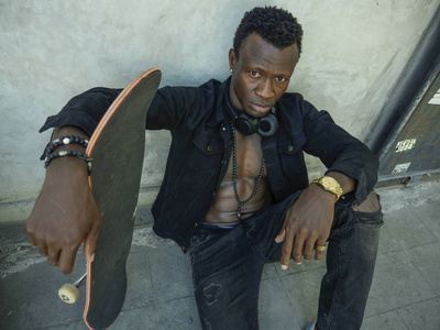 自信的时髦黑人非裔美国人靠在墙上举着滑板,是城市风格的坏蛋照片