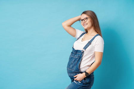 一位孕妇,戴着时髦的眼镜,抚摸着她的头发,在摄影棚的蓝色背景下微笑