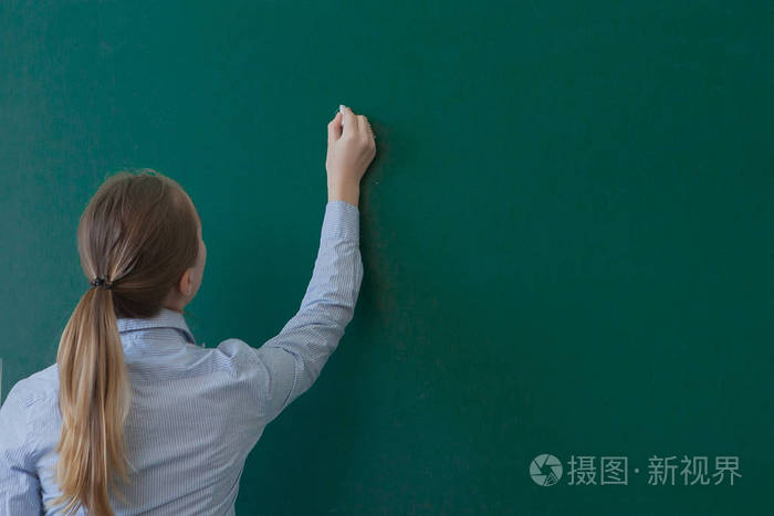 一个留着深色长发的学生或老师在空白的绿色黑板或黑板上写字的后视图